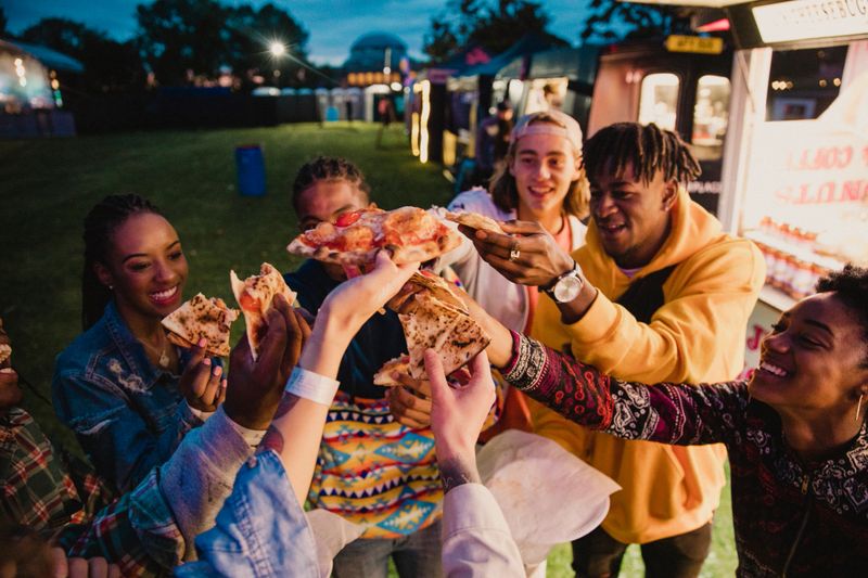 A young group of friends sharing and enjoying pizza at a music festival, making a celebratory toast with slices.