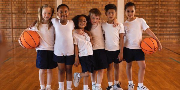 Six diverse kids in basketball uniforms posing with basketballs in a gym.