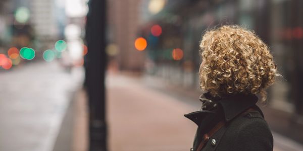 Person with curly hair stands on a city sidewalk looking away.