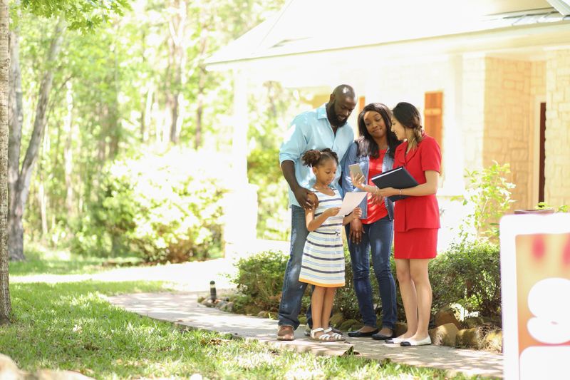 Latin descent Real Estate Agent shows African descent family a new home to purchase.  Mother, father and daughter.  Real estate sign.  Home in background.  Spring or summer season.