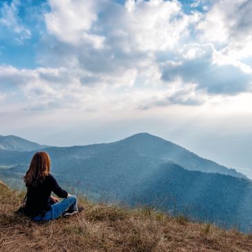 A person sitting on a hill, enjoying a scenic mountain view under a partly cloudy sky.
