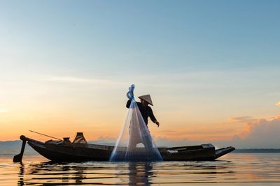 Fisherman casting a net from a boat at sunset on calm water.