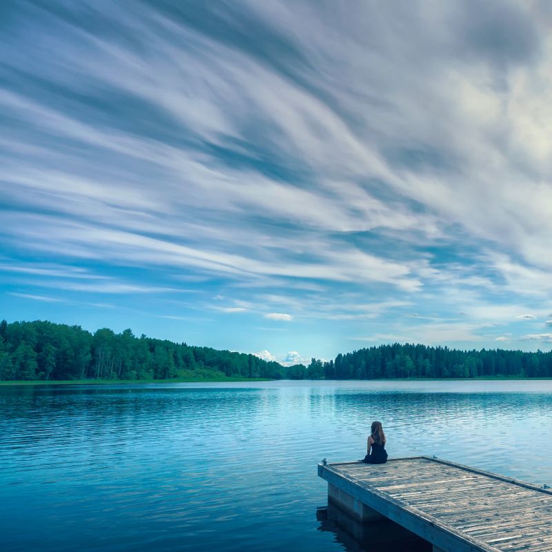 A woman sits on the edge of a jetty at peaceful lake with a dramatic sky.