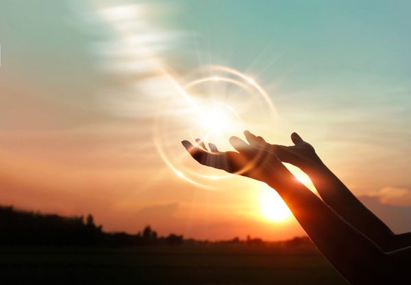 Hands reaching towards a glowing light at sunset.