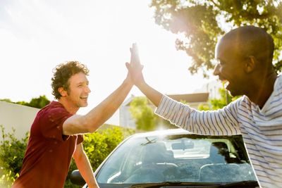 Two men happily high-five outdoors beside a car on a sunny day.