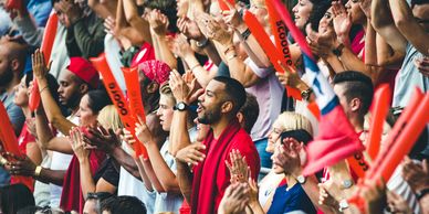 A diverse crowd of enthusiastic sports fans cheering and clapping with orange inflatable noisemakers.