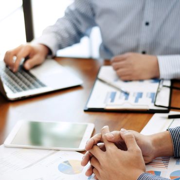 Two people working with charts, laptop, and tablet at a wooden table.