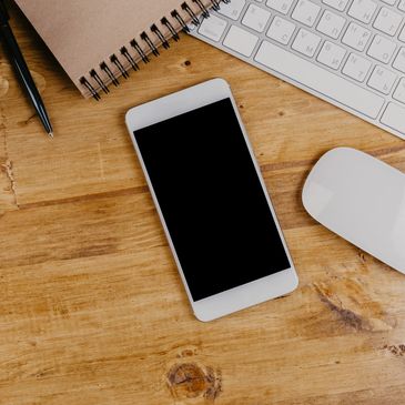 Smartphone, keyboard, mouse, pen, and notebook on wooden desk.