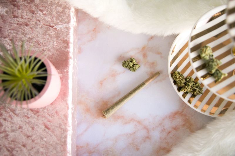 Top down shot of a marijuana joint and cannabis bud on pink marbled vanity. Vanity also contains a white and gold jewelry stand adorned with cannabis buds, a succulent in a pink pot, a pink velvet box, and white fur.