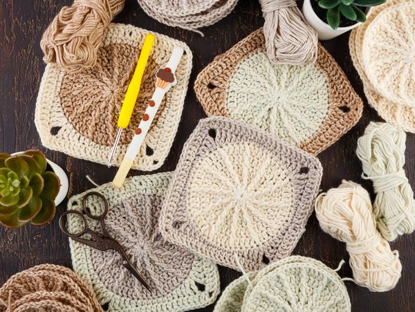 Crochet squares and circles with yarn, hooks, scissors, and succulents on a wooden table.