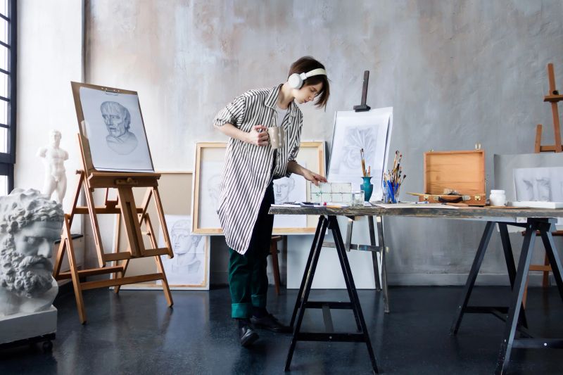 Modern freelancer artist sculptor woman in headphone creates new art masterpiece with cup of coffee at art workshop workplace studio exhibition with drawings, sketches and gypsum plaster sculptures.