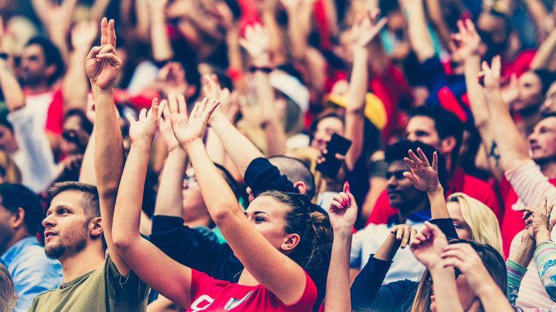 Crowd on a stadium cheering with hands up and waving with red balloons.