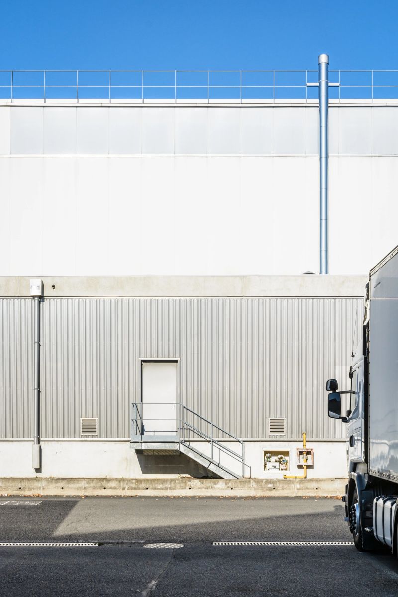 A white semitrailer truck parked opposite and perpendicularly to an industrial building with a pedestrian doorway at the top of a small staircase and covered with a grey corrugated iron siding.