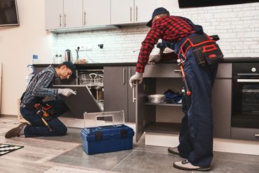 Two repairmen fixing kitchen cabinets and dishwasher with tools.