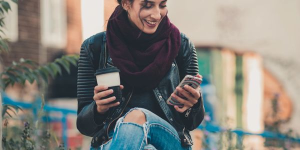 A woman smiling while looking at her phone and holding a coffee cup outdoors.