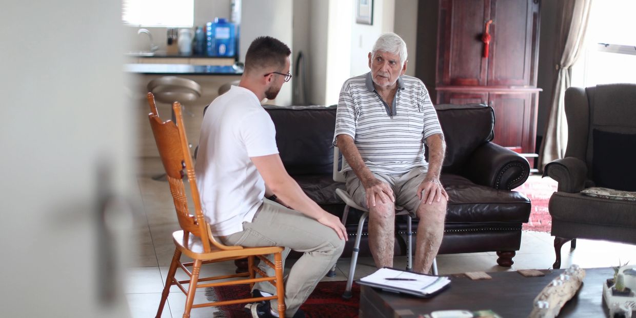 Younger man attentively talking to an elderly man sitting on a walker chair.