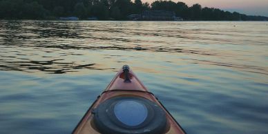 View from a kayak on calm water during sunset with trees in the distance.