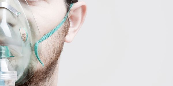 Close-up of a man wearing an oxygen mask against a plain background.