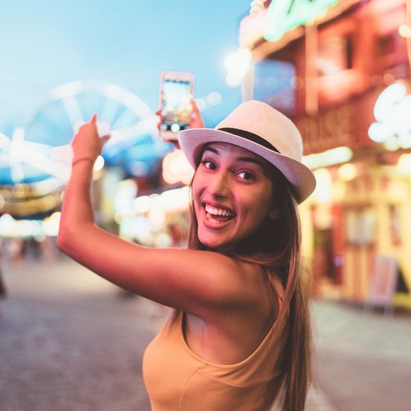 A joyful woman in a hat takes a selfie at a lively amusement park at dusk.
