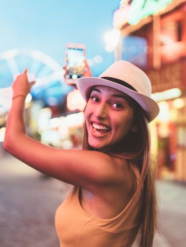 A joyful woman in a hat takes a selfie at a lively amusement park at dusk.