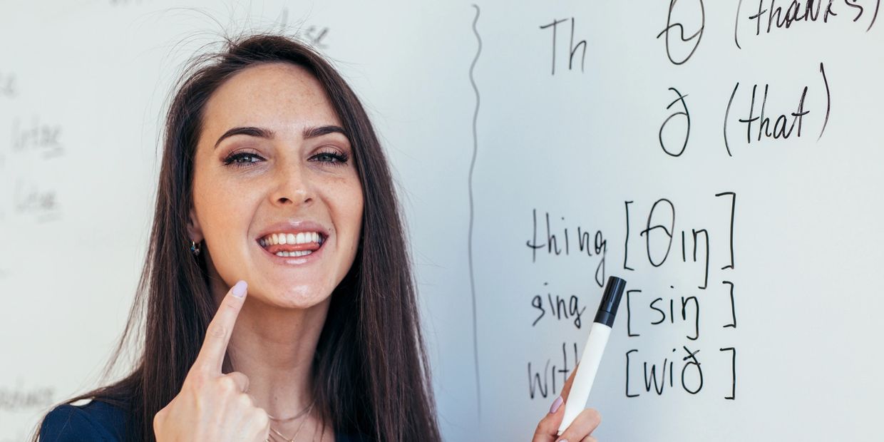 Accent Trainer explaining phonetic sounds on a board with /θ/, /ð/, & words like “thanks” & “that.”