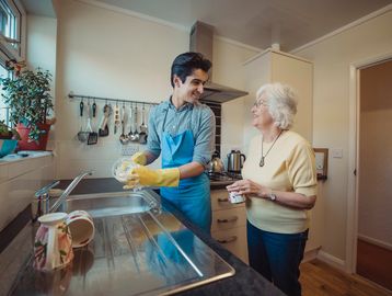 Young man washing dishes while chatting with elderly woman in kitchen.