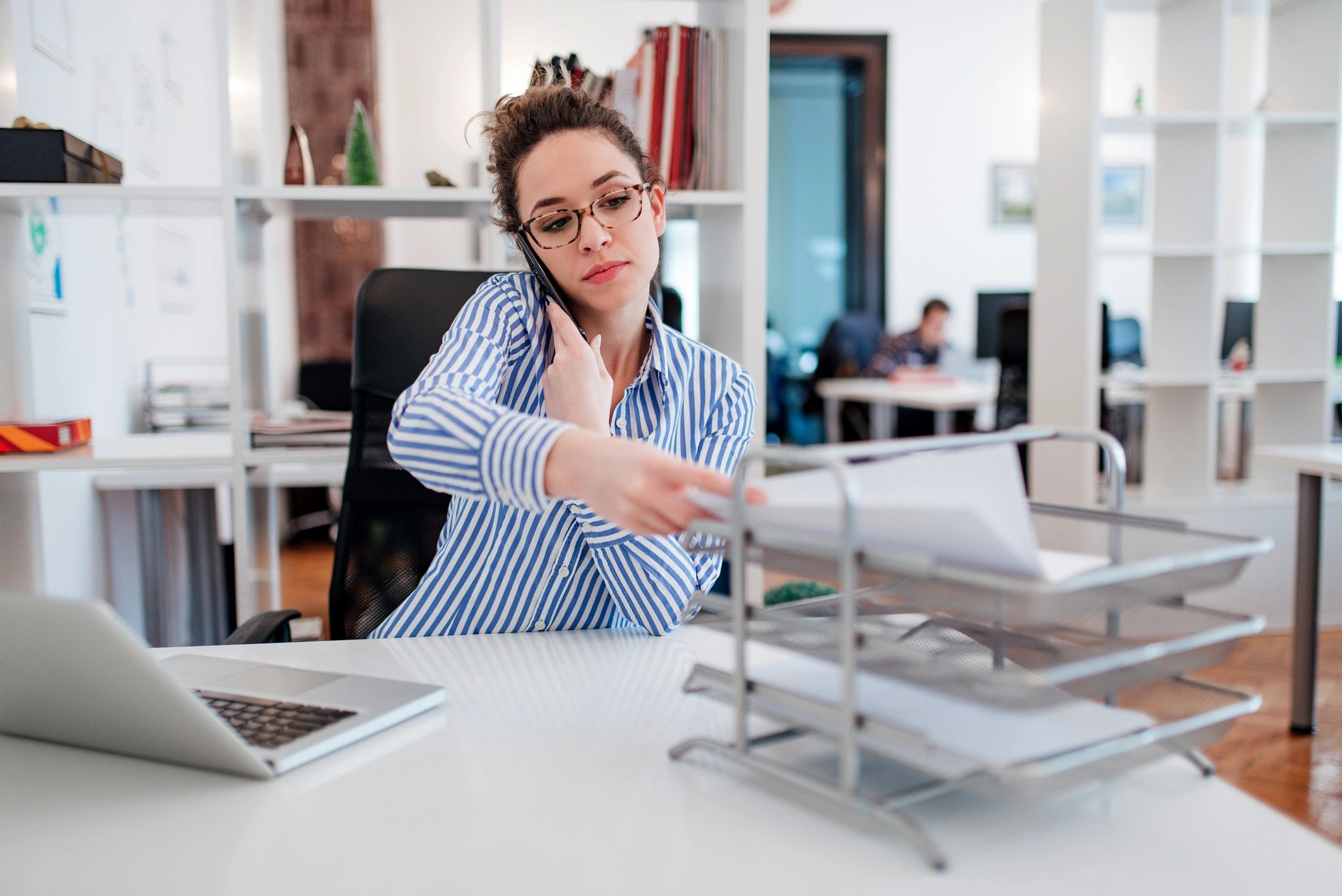 Woman multitasking at her office desk, talking on phone and organizing documents.