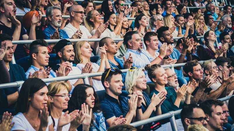 Large group of people sitting on stadium bleachers clapping.