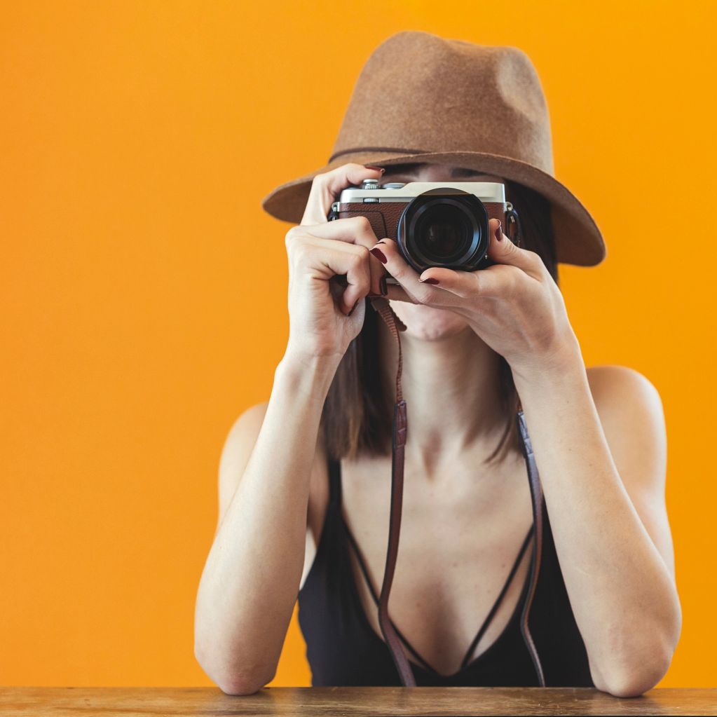 Woman in a brown hat taking a photo with a camera against an orange background.