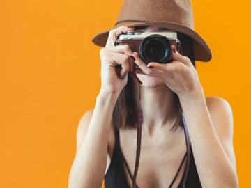 Person in a hat taking a photo with a camera against an orange background.