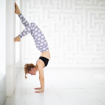 Woman practicing handstand yoga against a white wall in a bright room.
