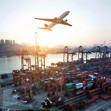 Airplane flying over a bustling cargo port at sunset.
