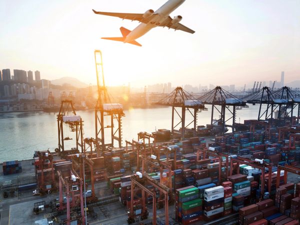 An airplane flies over a busy shipping port at sunset.