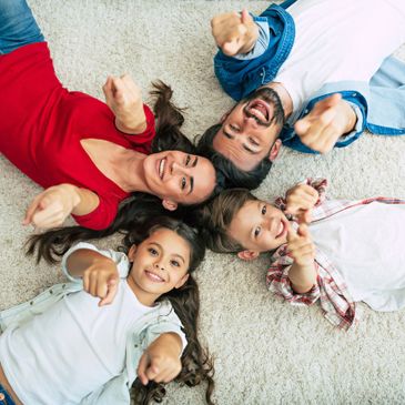 Happy family of four lying on carpet, smiling and pointing at camera.