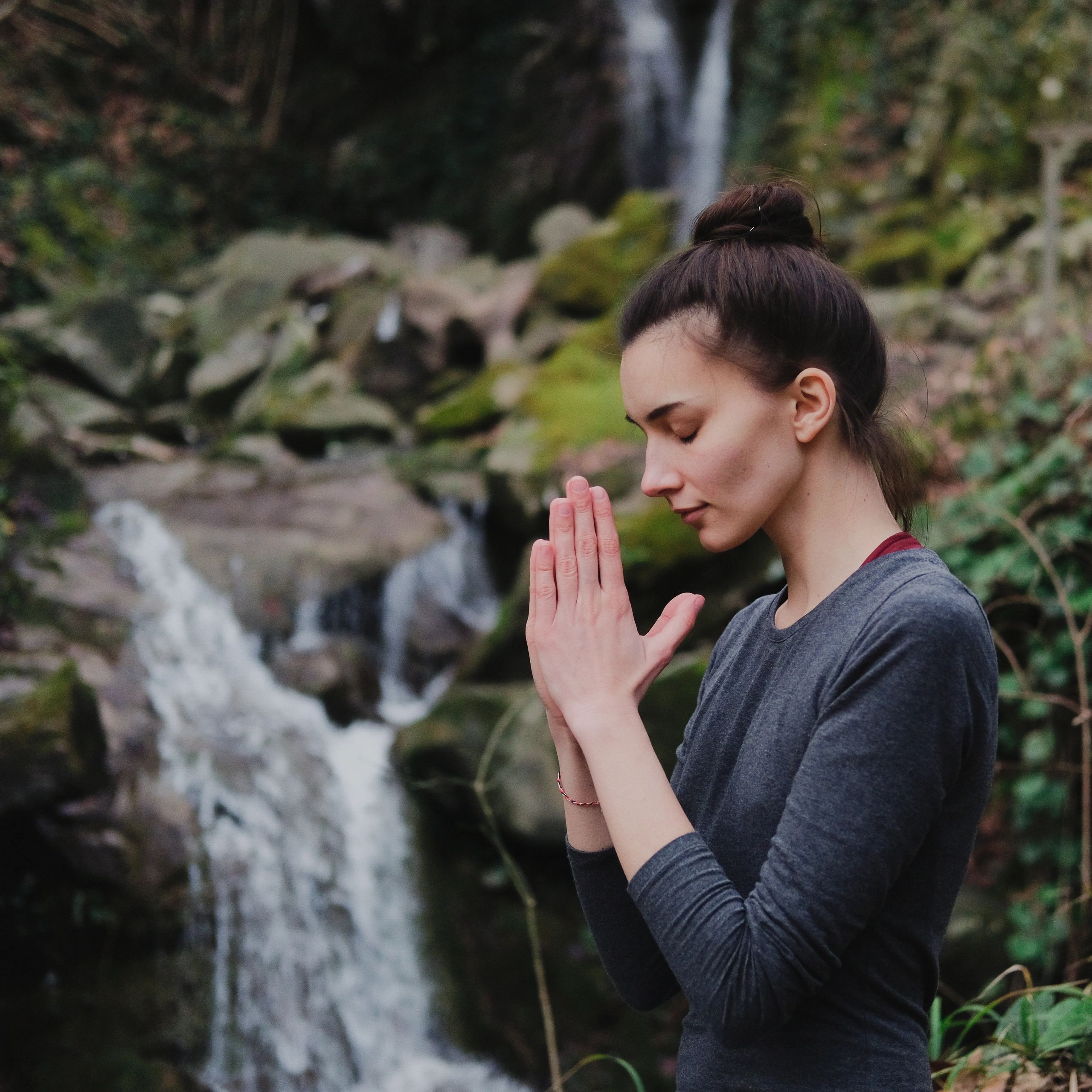 A woman meditates peacefully near a waterfall surrounded by nature.