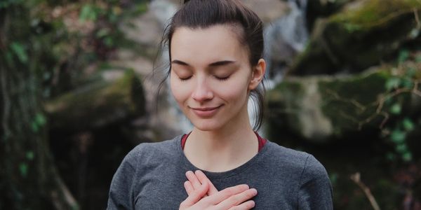 A woman with eyes closed, hands crossed over her chest, smiling peacefully outdoors.