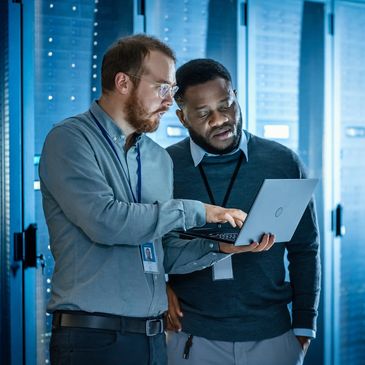 Two IT professionals discussing data on a laptop in a server room.