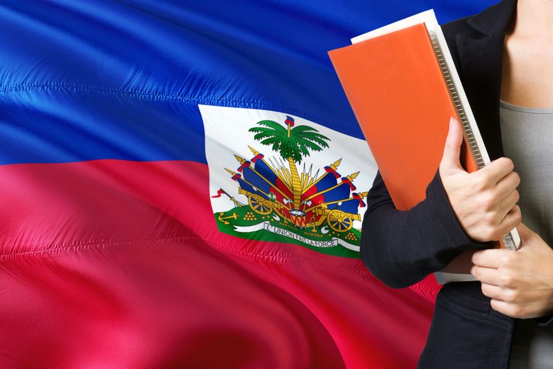 Learning Haitian language concept. Young woman standing with the Haiti flag in the background. Teacher holding books, orange blank book cover.