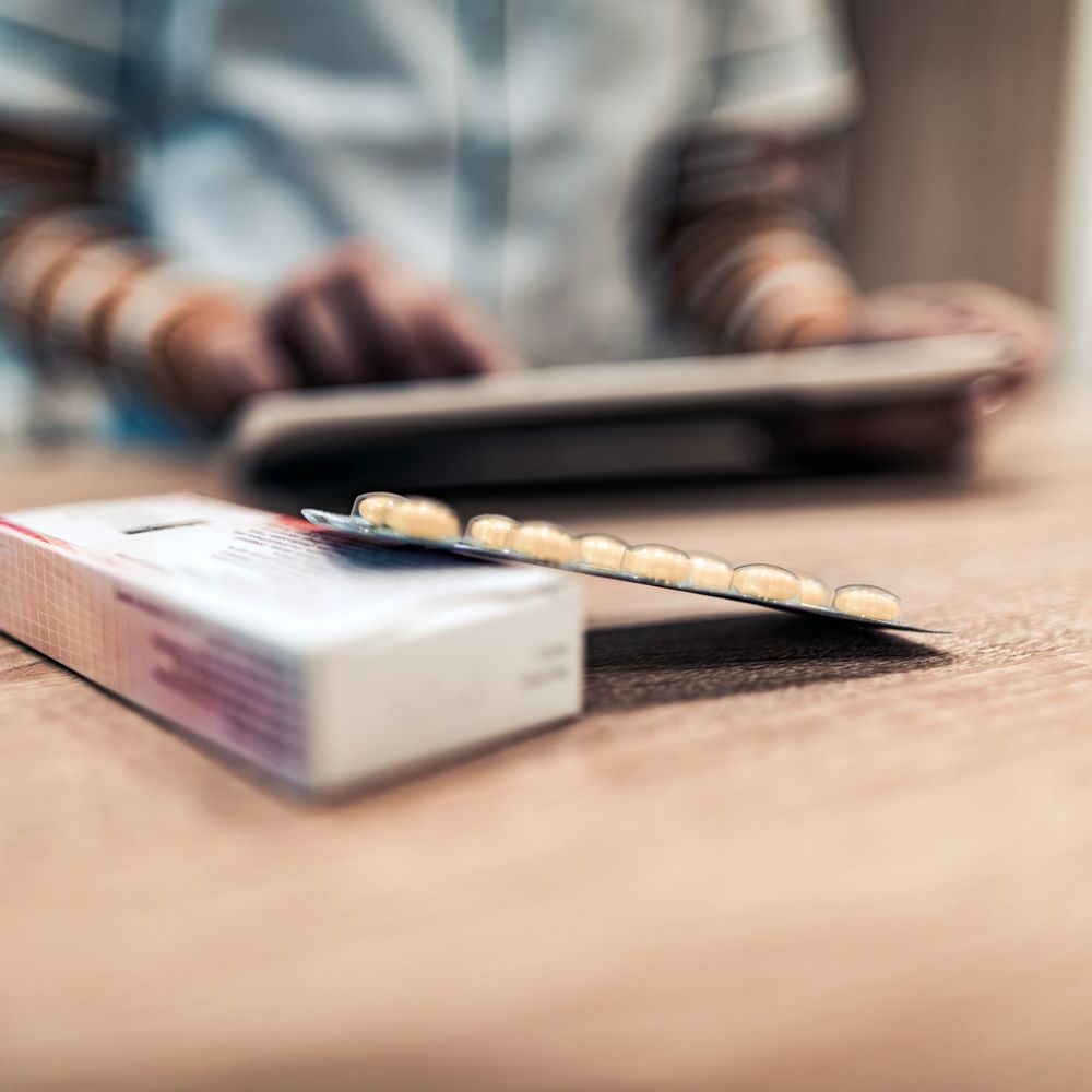 Blister pack of pills on a table with a person using a tablet in the background.
