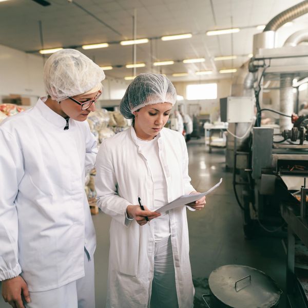 Two food factory workers inspecting production with safety gear and clipboard.