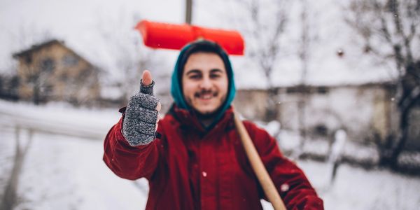 Smiling man in winter clothes giving a thumbs up while holding a snow shovel.
