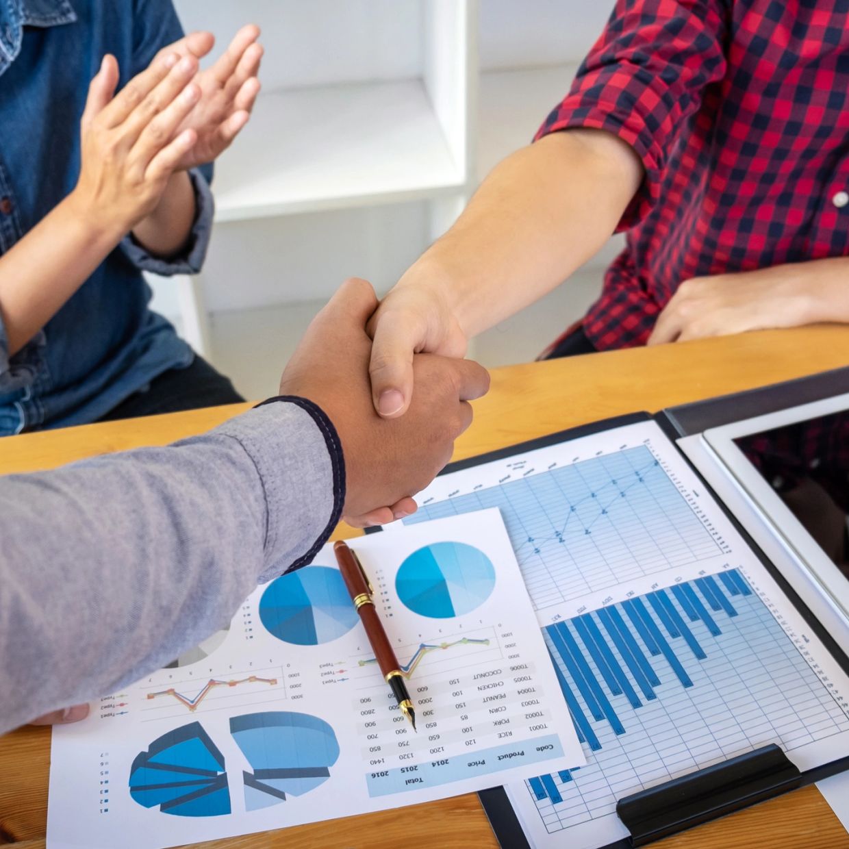 Handshake over business charts with another person clapping.