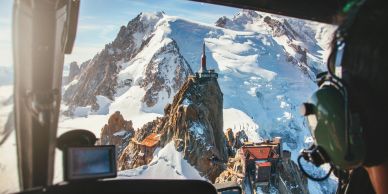 View of snowy mountains and a rocky peak from inside a helicopter cockpit.
