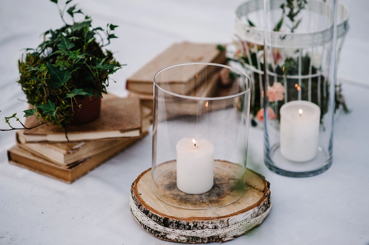 Lit candles in glass holders with rustic wood and plants on a white table.