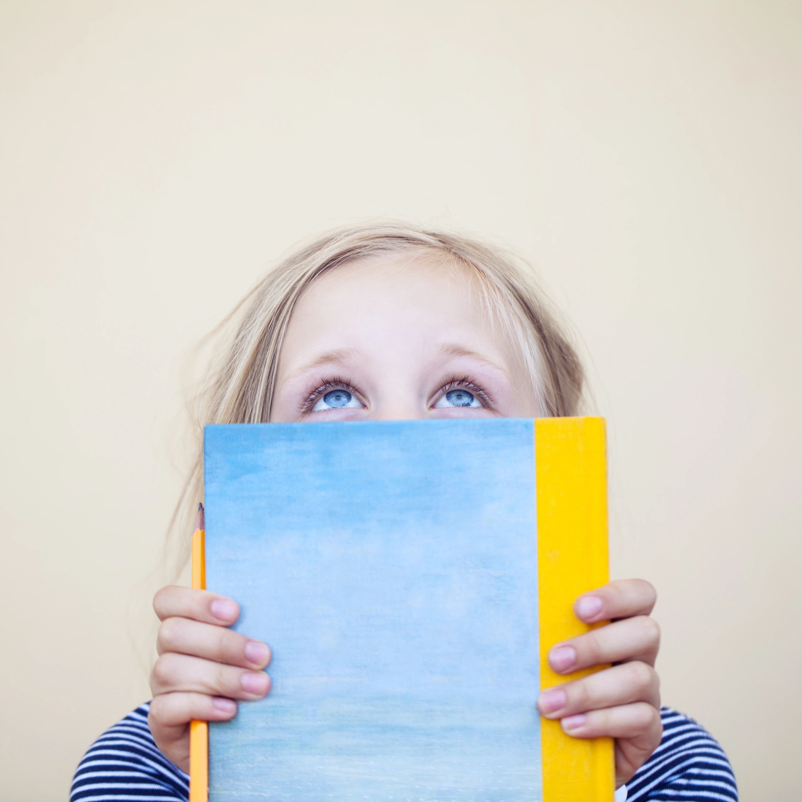 Child with blue eyes peeks over a blue and yellow book, holding a pencil.