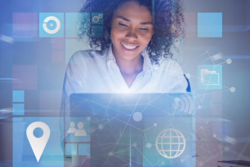 Cheerful African American businesswoman working with laptop in office with double exposure of business interface icons. Toned image