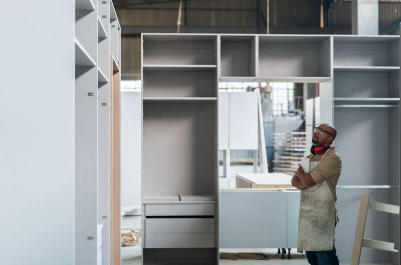 Confident craftsman looking at closet in factory. Bald worker is working at carpentry workshop. He is with digital tablet.