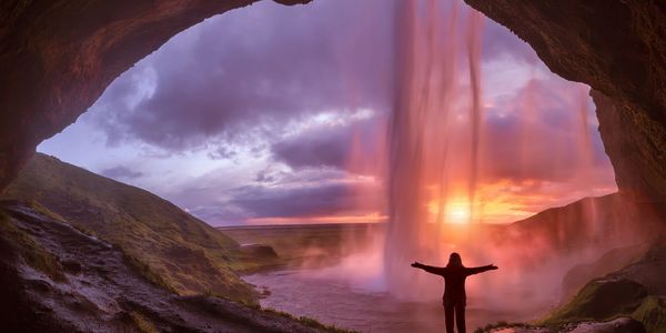 Person stands under a waterfall at sunset inside a cave.