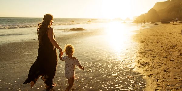 A woman and child hold hands walking along a beach at sunset.