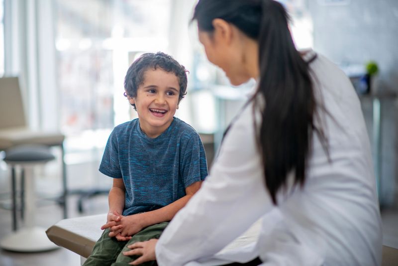 A young boy is indoors in a hospital room. His female doctor is giving him a checkup. The boy is smiling while the doctor checks his heart rate with a stethoscope.